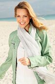 A blonde woman on a beach wearing a white blouse, a green leather jacket and a scarf