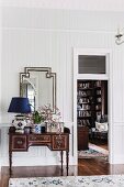 Table lamp with dark blue lampshade and vase of flowers on antique bureau against wood-clad wall and next to open door with view into library