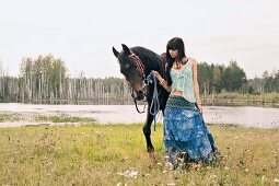 A young, dark-haired woman wearing hippie-style clothes leading a horse by a lake