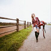 A long-haired blonde woman carrying a saddle and wearing a checked shirt and short shorts