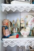 Doll, collectors' items and religious ornaments on shelf decorated with white lace trim
