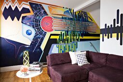 Bold, stylish mural behind aubergine corner sofa and vintage side table