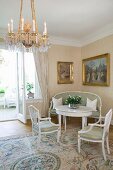 Delicate, white, antique-style table and chairs in corner below gilt-framed oil paintings; chandelier and silk rug in foreground