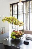 Fruit bowl and vases of flowers on black table in front of window with dark louvre blind