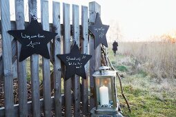 Star-shaped signs made from black-painted wooden hanging on a fence with a lantern with a candle with someone walking in the background