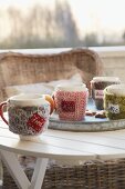 Mugs with knitted covers and name tags on a table outside