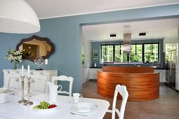 White dining set with neo-antique chairs with carved backrests; curved, wooden island counter in front of worksurface in kitchen area