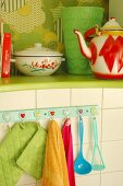 Kitchen utensils hanging from wall hooks under green-painted shelf holding enamel teapot and pan