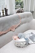 Korb mit Wollknäuel auf Sofa, über Lehne Tischtuch mit gehäkelter Borte in Rosa
