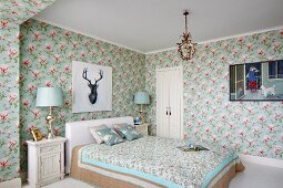 Double bed with white headboard and floral counterpane below portrait of stag on floral wallpaper in rustic bedroom