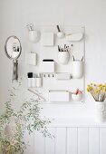 Vanity mirror and make-up in white organiser board above vase of yellow dried flowers on shelf