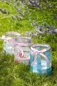 Drei Glaswindlichter mit romantischem Blumenmuster dekoriert und nostalgischem Flair im Gras neben Lavendelblüten im Garten
