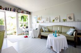 Open-plan living room with white wooden floor, round table, antique couch and drawings leaning against wall on top of wainscoting