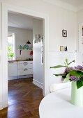 Vase of flowers on white table and view into modern kitchen through open doorway with continuous herringbone parquet flooring