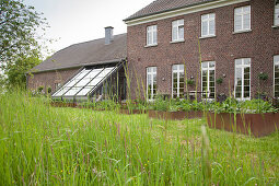 View from wild garden of raised beds with metal surrounds outside renovated, brick farmhouse