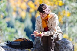 Frau sitzt mit Buch & Rucksack auf Felsvorsprung in herbstlicher Landschaft