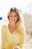 A young blonde woman on a beach wearing a white top and a pastel-yellow openwork jumper