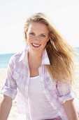 A young blonde woman on the beach wearing a white top and a purple and white checked blouse