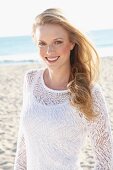 A young blonde woman on a beach wearing a white top and a transparent knitted jumper