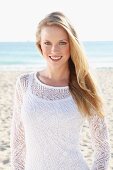 Junge blonde Frau in weißem Top und transparentem Strickpulli am Strand