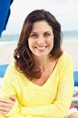 A brunette woman on a beach with her arms folded wearing a yellow knitted jumper