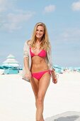 A young blonde woman on a beach wearing a pink bikini and a blouse