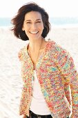 A brunette woman on a beach wearing a white top and colourful cardigan