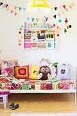 White, metal-grille bed with patchwork bedspread and colourful, patterned scatter cushions below bookshelf and colourful bunting on wall