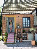 Woman in doorway of country house with brick façade holding flowerpot; striped runner on steps