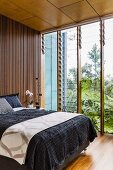 Double bed with black bedspread against wall with slatted wooden cladding next to glass wall with adjustable glass louvres