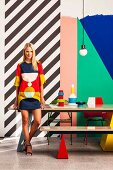 Junge Frau in Retro-Minikleid vor bunter grafischer Wandgestaltung und farbenfrohe Vasendekoration auf Esstisch