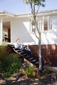 Couple sitting on rustic wooden steps without handrail outside white wooden house