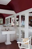 Vintage Standwaschbecken neben Einbauschrank mit Spiegelfüllung in Tür, im Vordergrund Armlehnstuhl in dunkelrot getöntem Bad