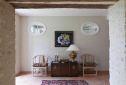 Open doorway in stone wall with view of wooden trunk between Scandinavian-style chairs below oval transom windows