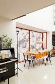 Offener Wohnraum, im Hintergrund Essplatz mit orangefarbener Tischdecke vor Fenster mit Rollos, seitlich schwarzes Klavier