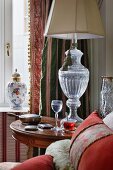 Lamp with urn-style glass base on antique side table, lidded china vase and classic, traditional curtains on window