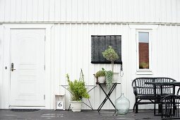 Haus mit weiss gestrichener Holzfassade, davor Grünpflanzen im Topf teilweise auf Tisch und schwarze Outdoormöbel auf Terrasse
