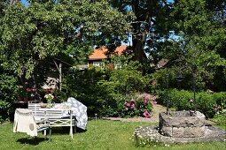 Gartenplatz mit weissen Sitzmöbeln in sonnenbeschienenem Garten, seitlich alter Ziehbrunnen aus Stein
