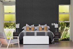 Doppelbett und weiße Truhe in modernem Schlafzimmer mit dunkelgrau getönter Wand, seitlich raumhohe Fenster mit Gartenblick
