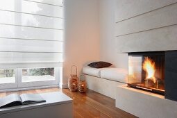 Fireplace next to bench with white seat cushions and candle lanterns on parquet floor in corner in front of French windows with closed roller blinds