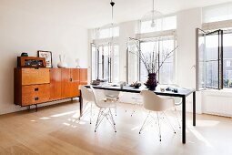 Modern table, white classic shell chairs, transparent pendant lamps and sideboard in retro interior with large windows