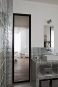 Full-length mirror next to masonry washstand clad in shiny silver mosaic tiled in bathroom