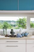 Ausschnitt einer Küchenzeile, Holz Arbeitsplatte auf weißem Unterschrank vor Fenster, oberhalb Hängeschrank in Blau