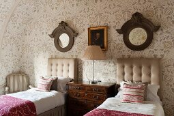 Twin beds with button-tufted headboards, antique chest of drawers, round mirrors with wooden frames and floral wallpaper in attic bedroom in English manor house