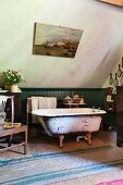 Free-standing vintage bathtub against green-painted wainscoting on sisal rug in bathroom with sloping ceiling