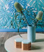Exotic flowering plant in light blue base on table, wallpaper with leaf motif behind it