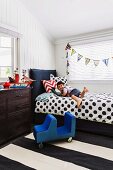 Boy lying on bed with black and white polka-dot bedspread below bunting and next to chest of drawers in child's bedroom