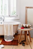 Large seashell on rustic wooden stool next to free-standing bathtub below window in bathroom with maritime atmosphere