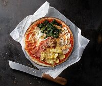 Pizza mit Spinat, Champignons, Artischocken, Parmaschinken, Zwiebeln und Thunfisch auf Papier mit Messer