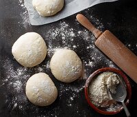 Four pizza dough balls on a baking tray with flour and a rolling pin